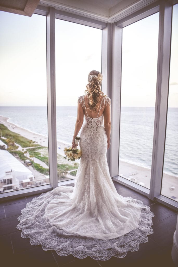 The bride looks our her ocean front suite at Eden Roc hotel in Miami Beach in her Eve of Milady wedding dress with beaded sleeves and long lace train