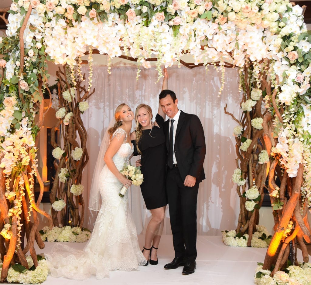 The bride and groom pose with the best wedding planner in South Florida under their gorgeous chuppah filled with greenery and orchids at The Bath Club