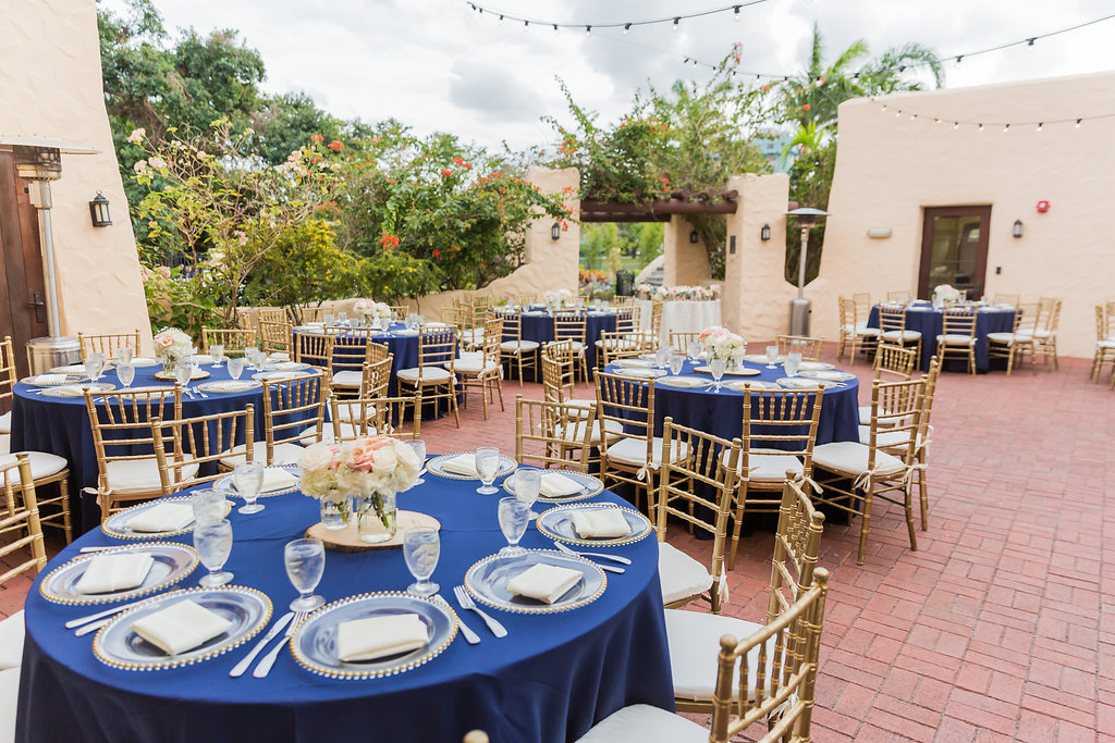 The Curtiss Mansion wedding had dinner in the courtyard with navy blue linens, gold beaded charger plates, gold chiavari chairs, and ivory seat cushions. Bistro lights hung from above