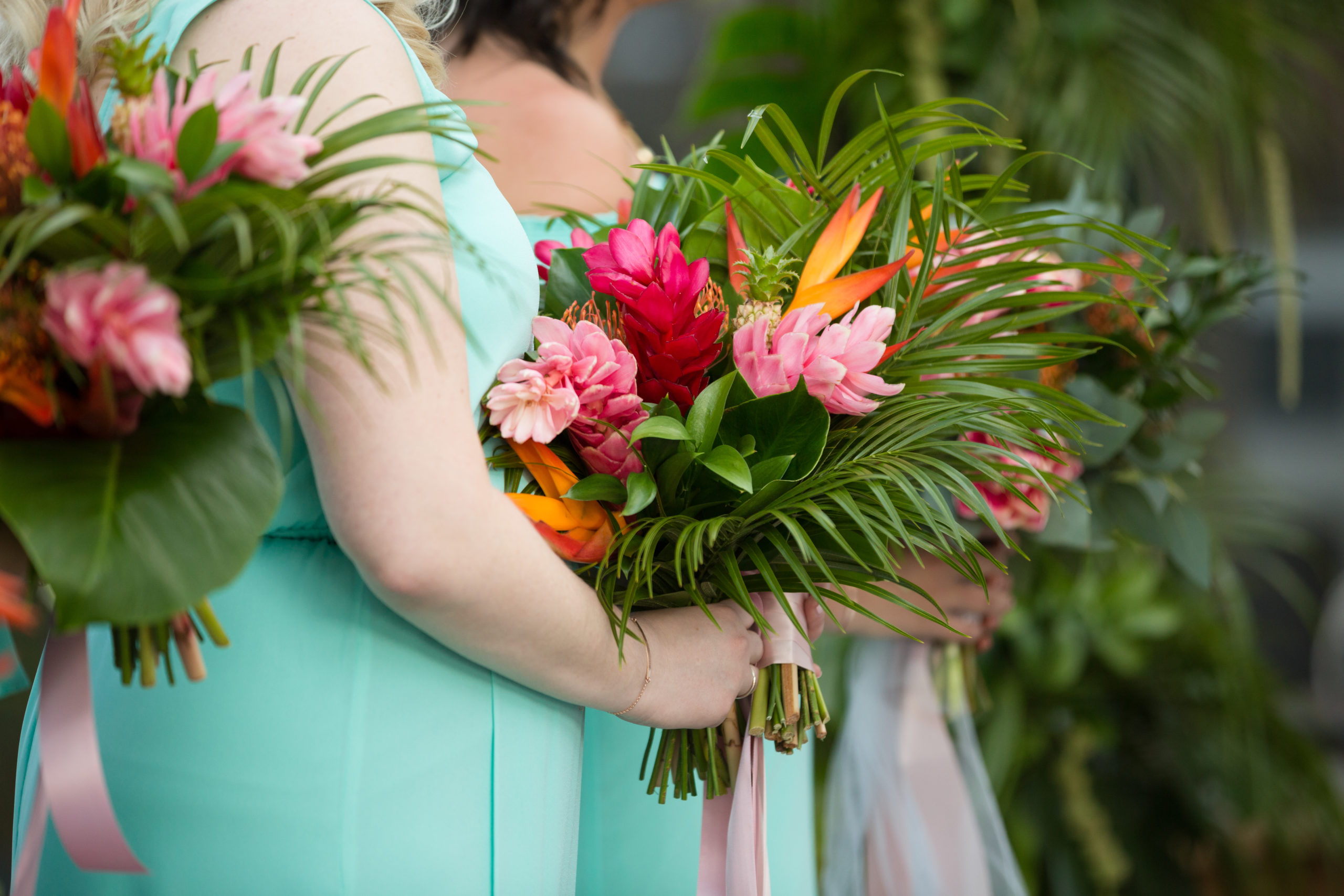The bridesmaids carry tropical bouquets filled with monstera leaves, banana leaves, protea, birds of paradise, pineapples, and other tropical flowers