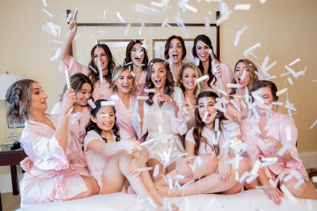 the bridesmaids sit on a bed shaking confetti in blush robes