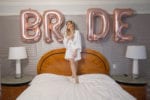 bride stands on her bed in a white robe with a champagne flute with BRIDE balloons at four seasons Miami