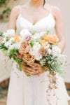 the bride holds a loose and flowing bouquet with peach roses, sahara roses, mauve roses, white and ivory flowers, and loose hanging greenery