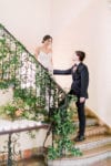 the bride and groom stand on the villa woodbine staircase that has been decorated in loose greenery