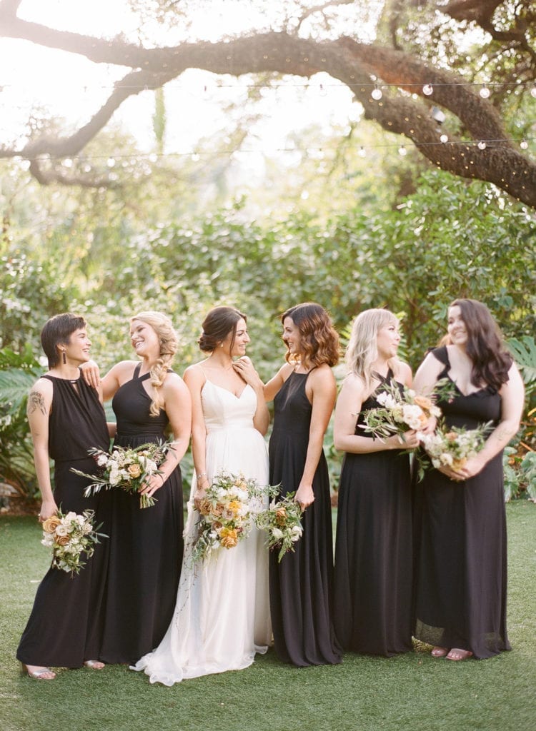 the bridesmaids are wearing varying style black dresses with loose bouquets in their hands while they smile and laugh with the bride