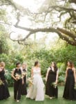 bride walks under a tree with her bridesmaids wearing all black