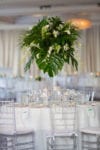 a tall tropical centerpiece with monstera leaves and hanging orchids on a table with clear chiavari chairs