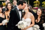 bride and groom share a sweet kiss as their wedding party smiles and hugs them in a wedding designed by Oh My Occasions