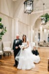 bride and her father stand arm in arm as they get ready to walk down the aisle and Oh My Occasions flares out her wedding dress train