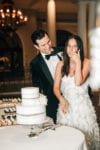 bride licks her finger as the groom smiles behind her while cutting their wedding cake