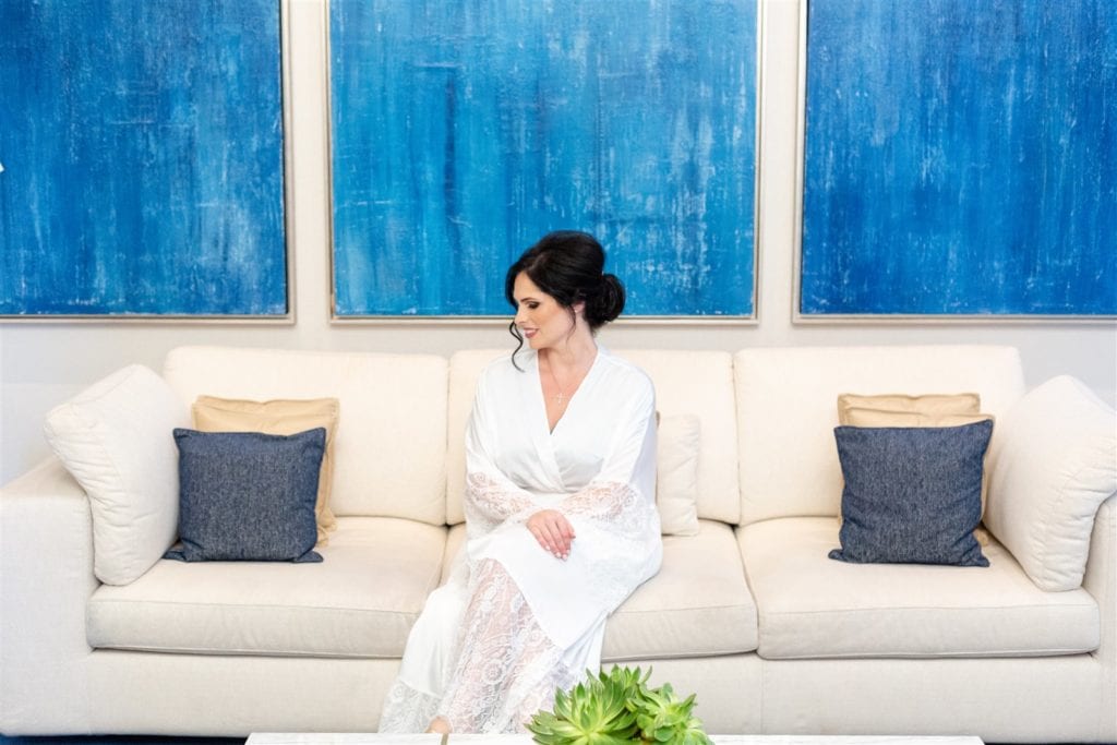 The bride in her white robe sitting down on a couch at The Conrad Fort Lauderdale Beach, wedding planned by Oh My Occasions