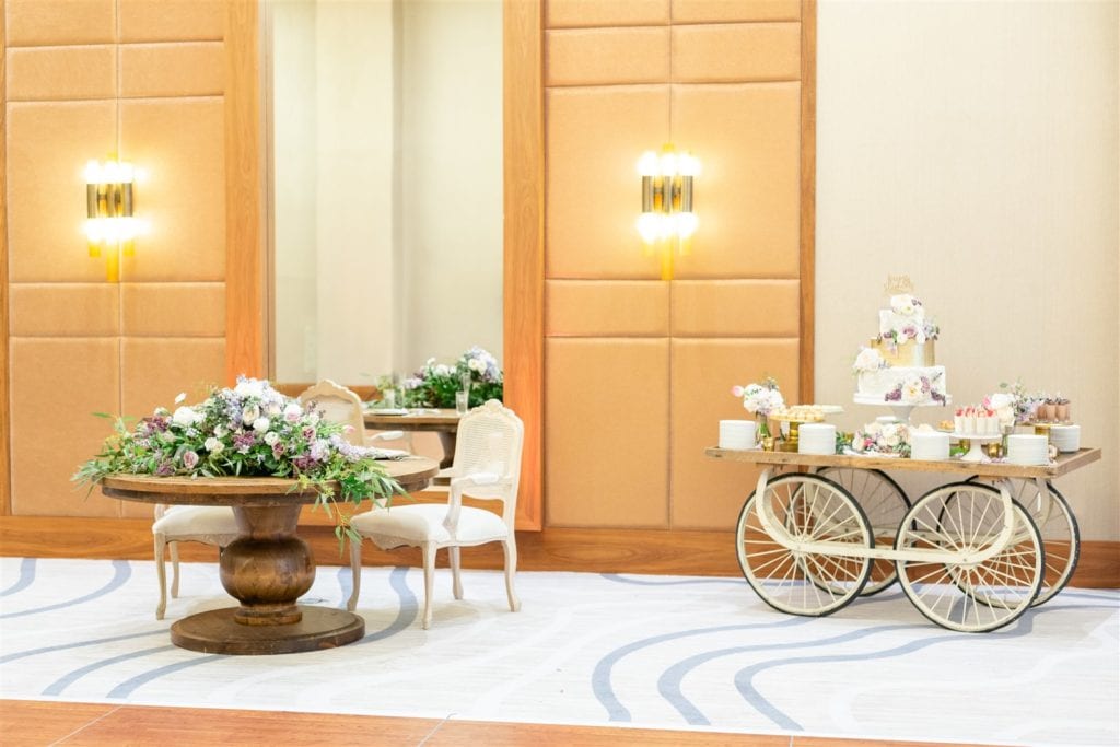 The reception venue with a strolling cart with the wedding cake on top with a round wooden table adorned with flowers at The Conrad Fort Lauderdale Beach, wedding planned by Oh My Occasions
