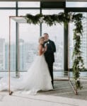 bride and groom stand smiling under their copper chuppah with greenery