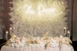 sweetheart table with ivory textured linen and large feather wall backdrop with neon sign