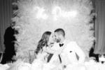 bride and groom kiss in front of their feather wall wedding backdrop with neon sign