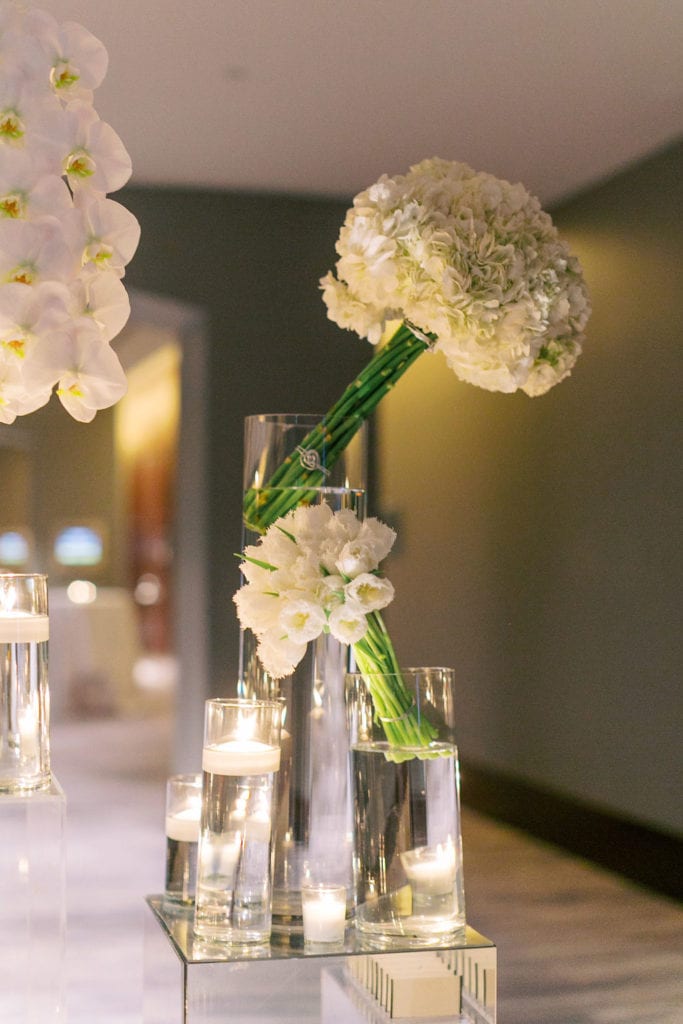 A closeup of the name card arrangement decorated with bouquets of hydrangeas and orchids in a tall pillar vase glasses filled with water at The Mandarin Oriental, wedding planned by Oh My Occasions