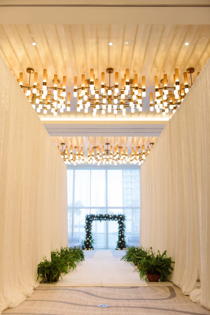 Beautiful wedding aisle with white draping, greenery along the aisle, and green arch full of flowers at the Conrad Fort Lauderdale Beach, wedding planned by Oh My Occasions