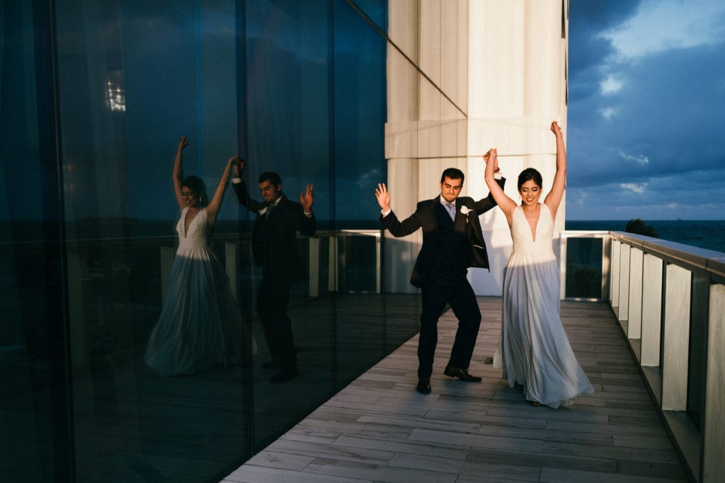 The bride and groom dancing and waving their hands in the air outside the balcony at the Conrad Fort Lauderdale Beach, wedding planned by Oh My Occasions