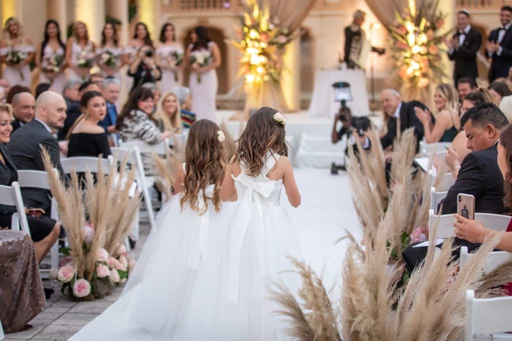 Two flower girls wearing matching white dresses walk down the aisle showing the back at The Baltimore Hotel in Miami Coral Gables, wedding planned by Oh My Occasions
