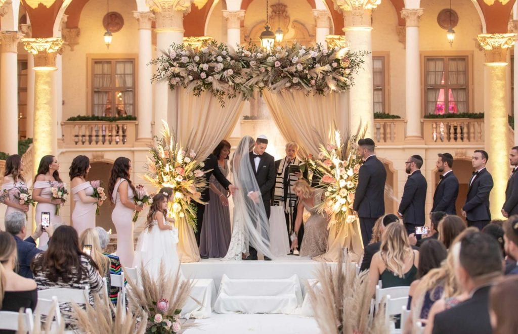 The wedding couple and parents stand under the chuppah with rabbi while wedding party stand beside them at The Baltimore Hotel in Miami Coral Gables, wedding planned by Oh My Occasions