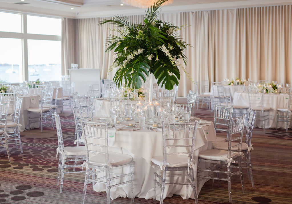 A round table with white table linens, clear Chiavari chairs and a tall palm leave centerpiece at The Rusty Pelican, planned by Oh My Occasions 