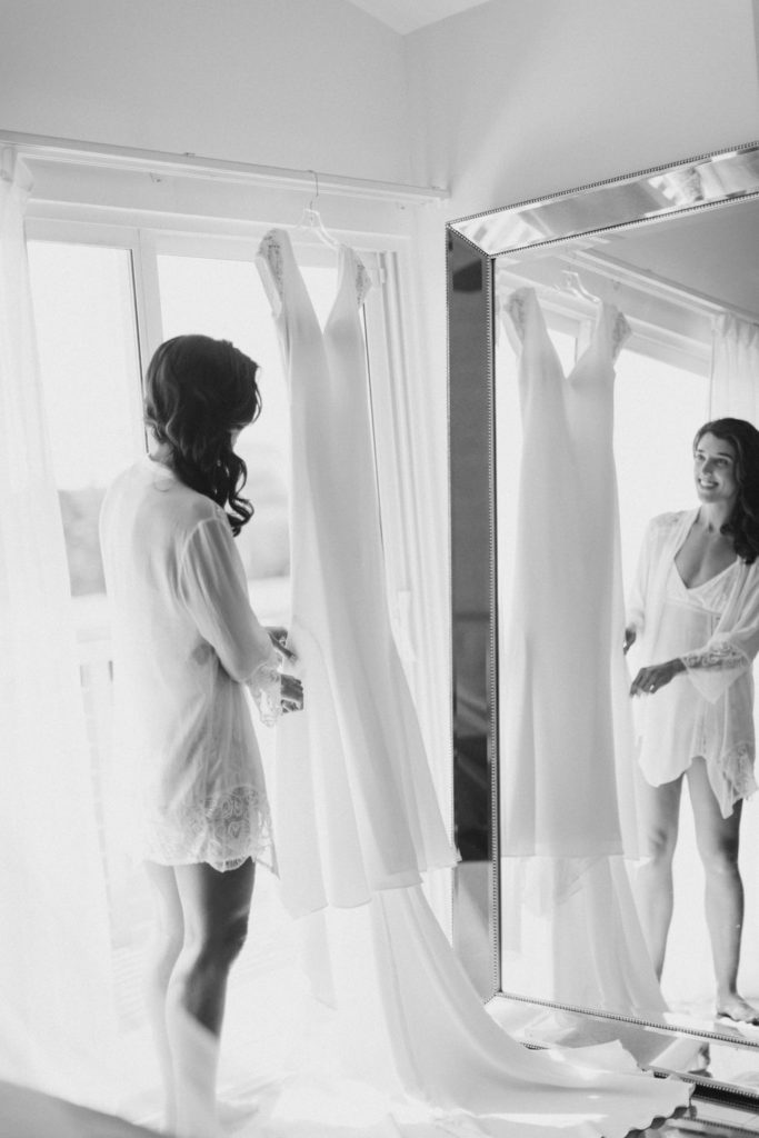 Black and white photo of bride admiring her hung wedding gown in her getting ready outfit.