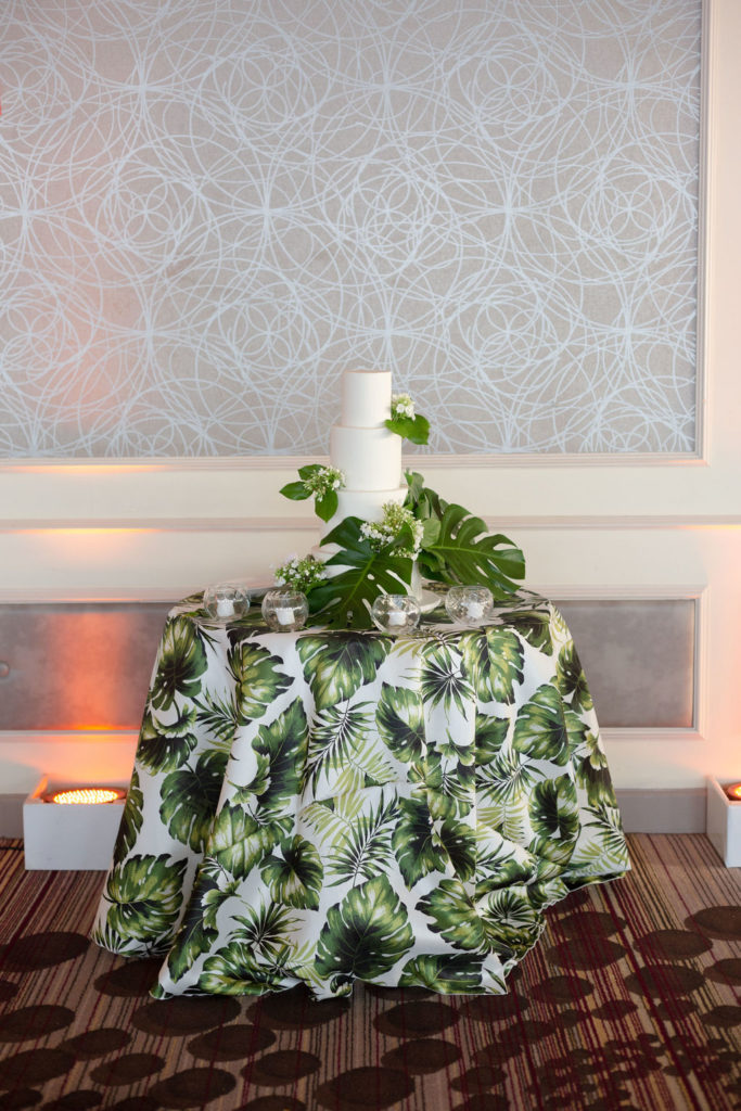 Four-layer white wedding cake with palm leaves on top of a tropical monstera table linen at The Rusty Pelican, planned by Oh My Occasions 