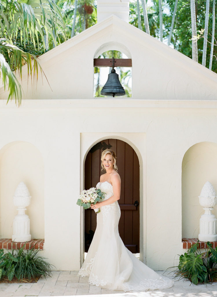 Bride stands in front of door and smiles at the camera with her bouquet wearing a strapless gown.
