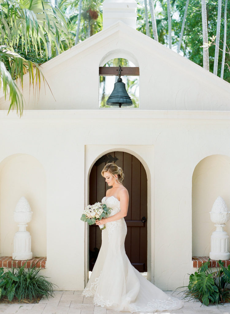 Bride stands in front of door and looks down at her bouquet wearing a strapless gown.