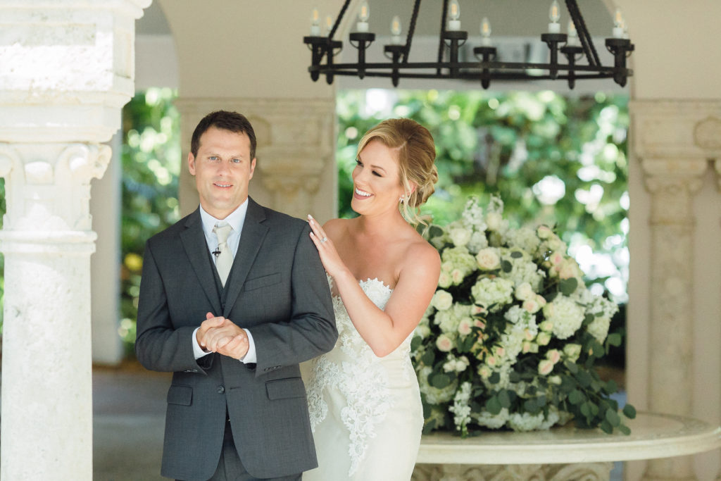 Bride stands behind groom and smiles at him while grooms smiles at camera for their first looks at Fisher Island Club.