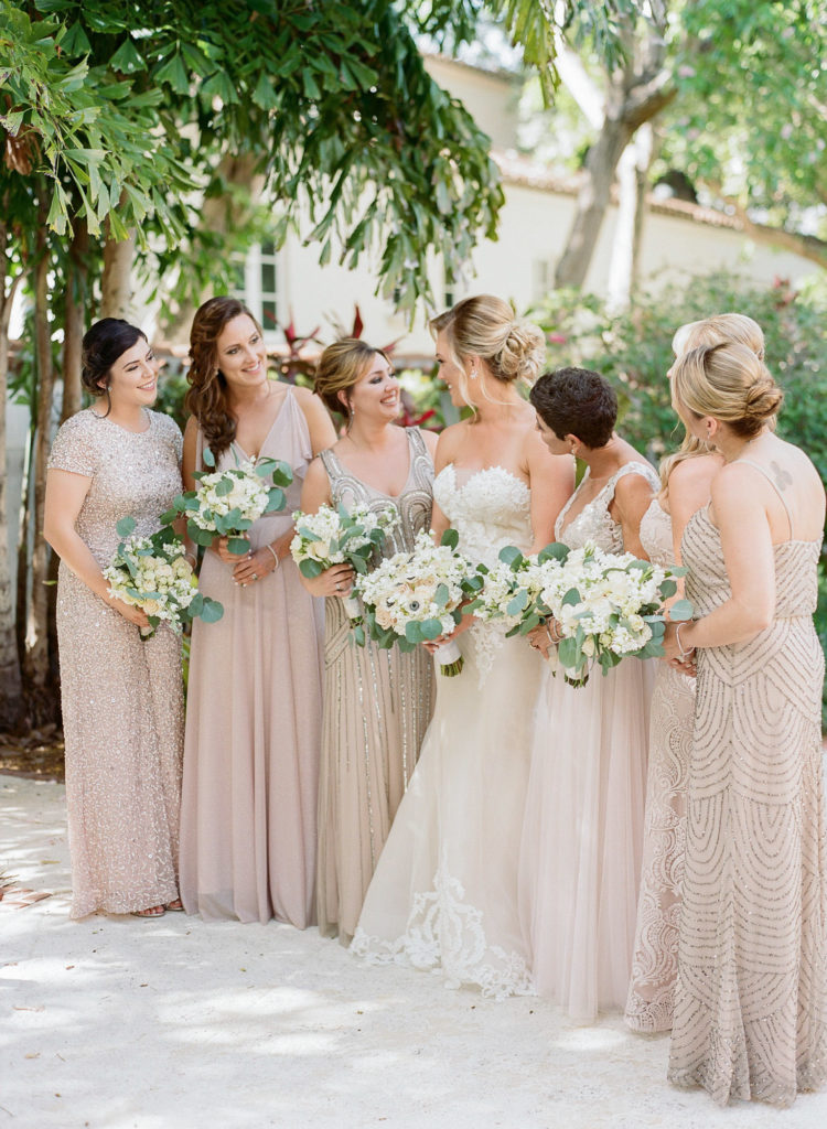 Side photo of bride standing with her bridal party looking at each other wearing blush mix-matched gowns and holding their bouquets.