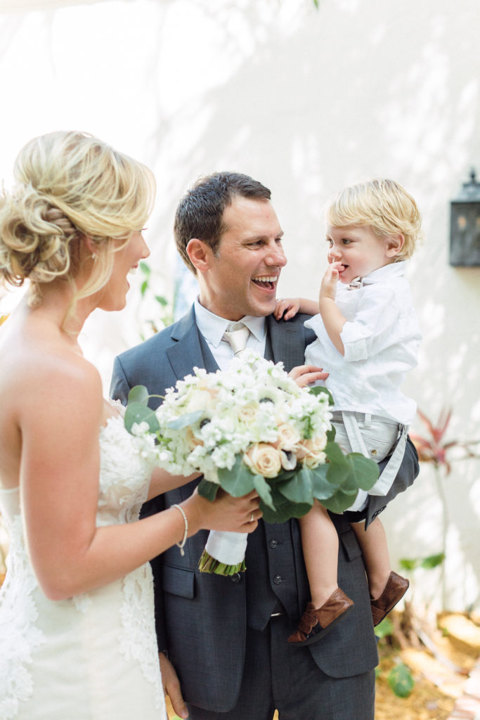 Groom carries couple's son while bride smiles at them holding her bouquet.