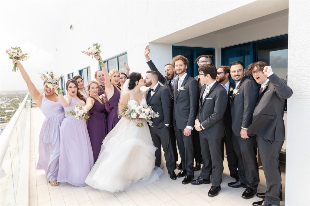The bride and groom kissing with the groomsmen and bridesmaids behind them cheering at The Conrad Fort Lauderdale Beach, wedding planned by Oh My Occasions