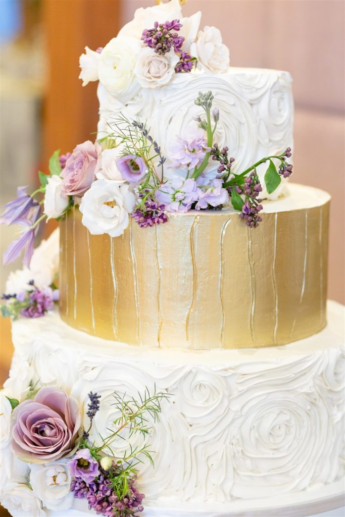 A three-tiered wedding cake with a gold layer adorned with purple flowers at The Conrad Fort Lauderdale Beach, wedding planned by Oh My Occasions