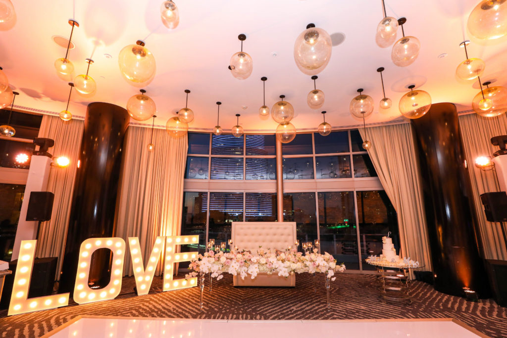 A beautiful overview of the specialty sweetheart table adorned with roses, alternating bubble lights, and a LOVE marquee sign