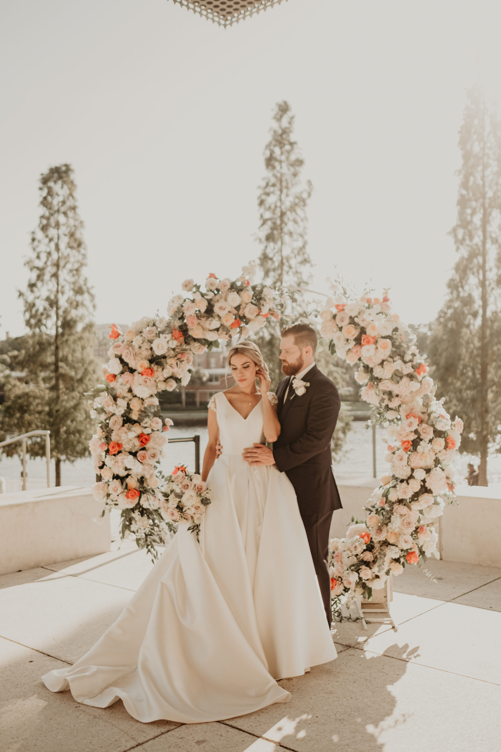 A lovely shot of the bride and groom embracing with a round floral arch adorned with pink and white flowers at The Tampa Museum of Art, wedding planned by Oh My Occasions