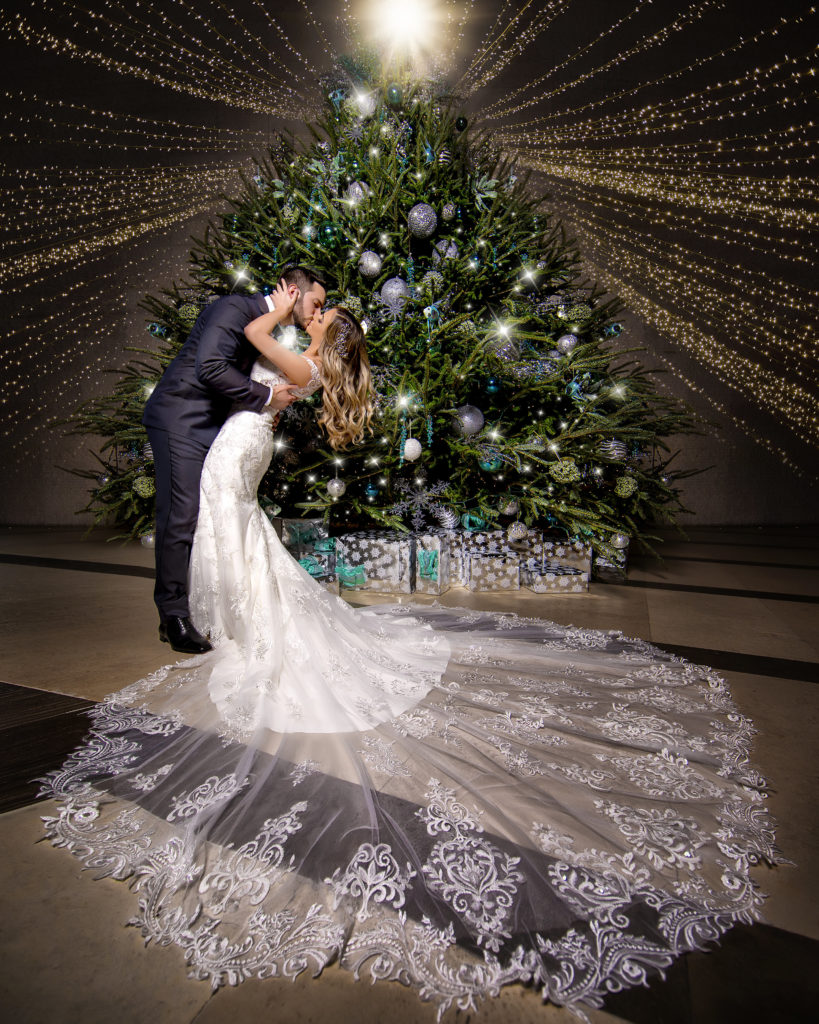 The bride and groom kissing in front of a beautifully lit Christmas tree with lights hanging at the Kimpton EPIC Hotel, planned by Oh My Occasions
