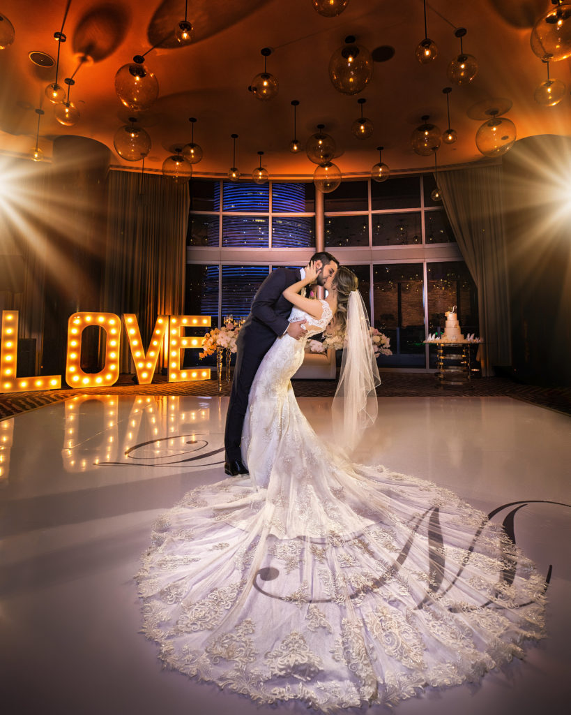 A stunning shot of the bride and groom kissing on the custom wrapped dance floor with the LOVE marquee sign in the back at the Kimpton EPIC Hotel, planned by Oh My Occasions