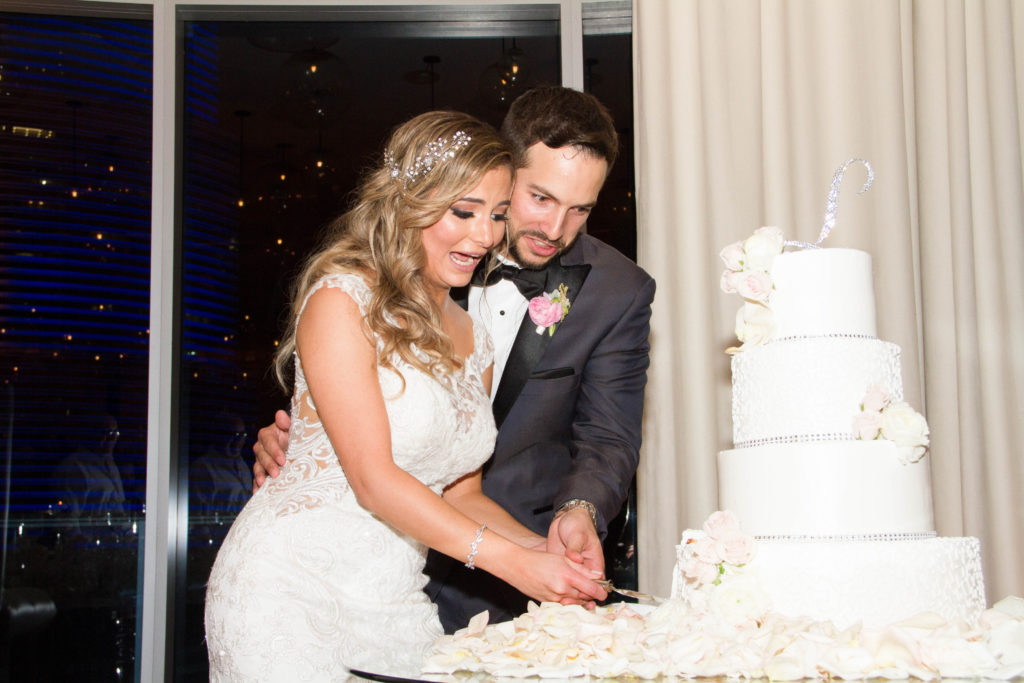 The bride and grooms slicing their four-layered white cake with roses and a bedazzled belt and ripple tiers at the Kimpton EPIC Hotel, planned by Oh My Occasions