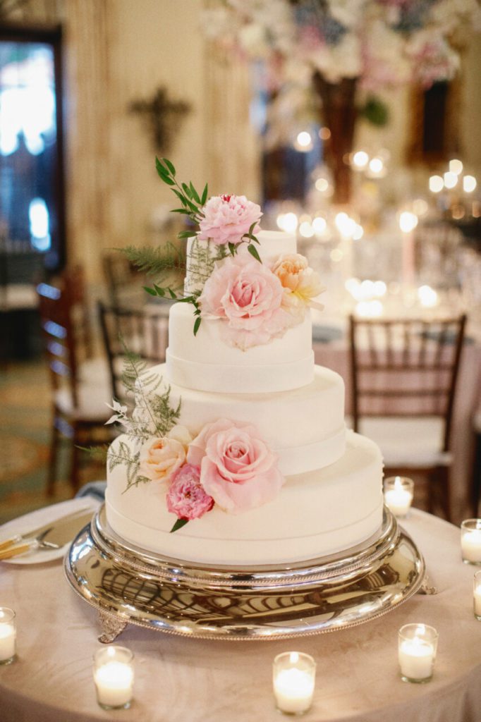 A stunning four-layered cake with white frosting, adorned with delicate light pink roses and a touch of greenery for a natural, elegant look at Indian Creek Country Club planned by Oh My Occasions.