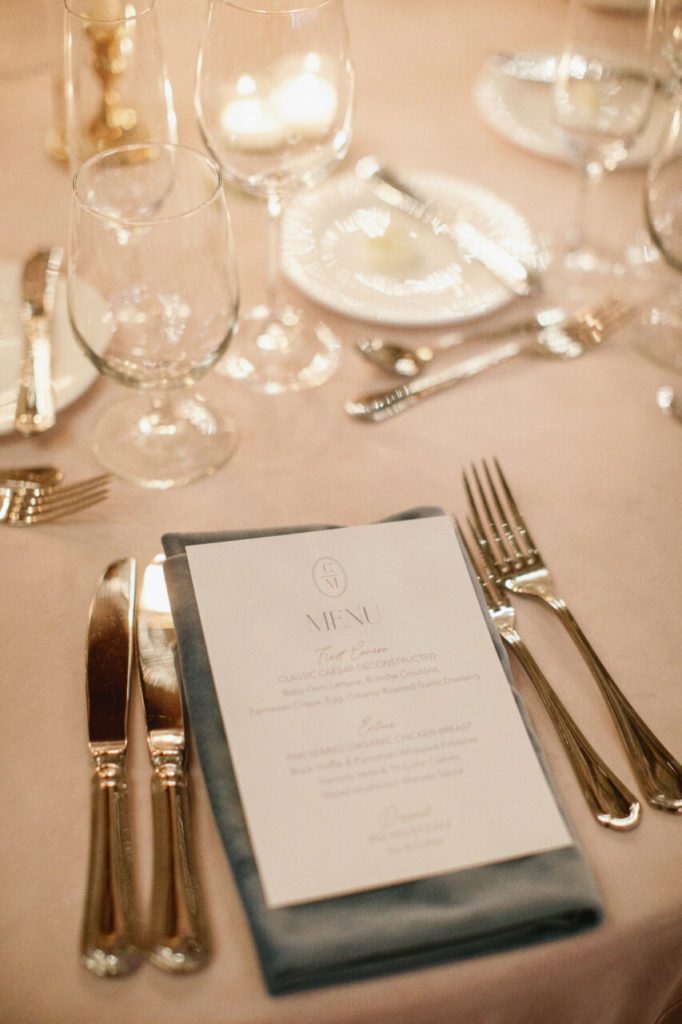 White menu cards with silver cutlery and clear glass cups, sitting on top of a soft pink table linen with dim candle light at Indian Creek Country Club planned by Oh My Occasions.