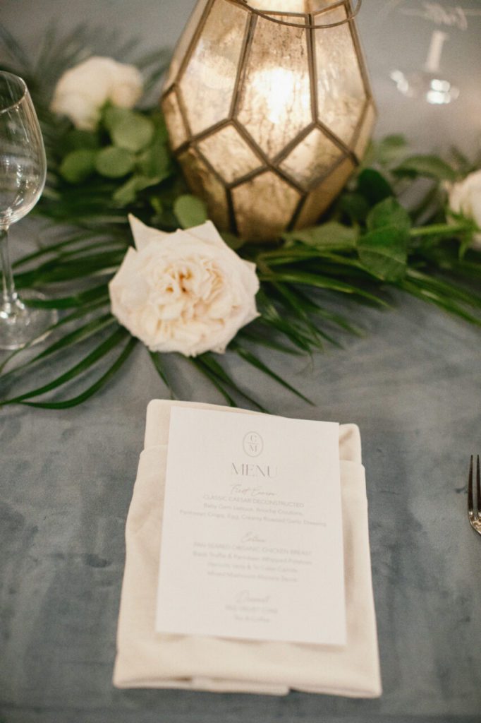 A dinner menu place card on top of a velvet blue table linen decorated with white roses and geometric terrarium vessels at Indian Creek Country Club planned by Oh My Occasions.