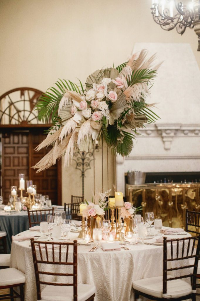 An exquisite reception table adorned with a stunning centerpiece featuring a beautiful arrangement of pampas grass, white hanging amaranthus, and delicate white and light pink roses. The gold vintage candles add a touch of elegance and romance to the table at Indian Creek Country Club planned by Oh My Occasions.