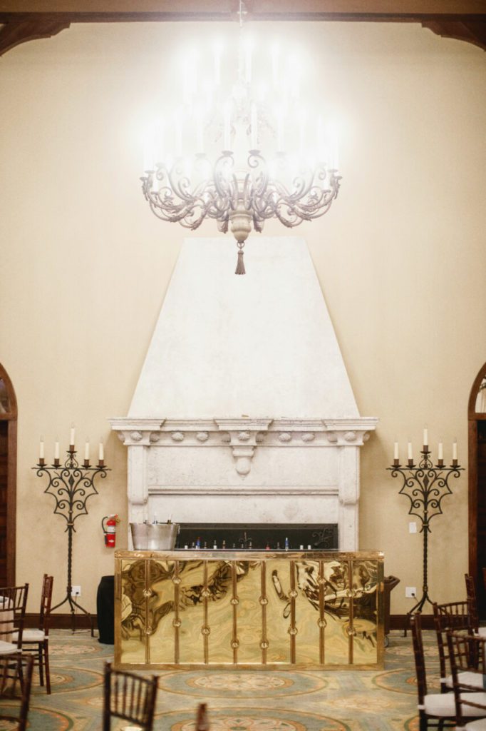 A beautiful gold reflective bar table sits in front of a large white chimney in between two French Candelabra Lamps at Indian Creek Country Club planned by Oh My Occasions.
