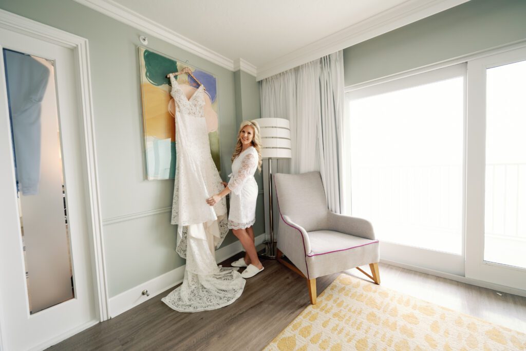 a bride stands with her hanging dress in the suite of La Playa Golf and Beach Resort in Naples Florida